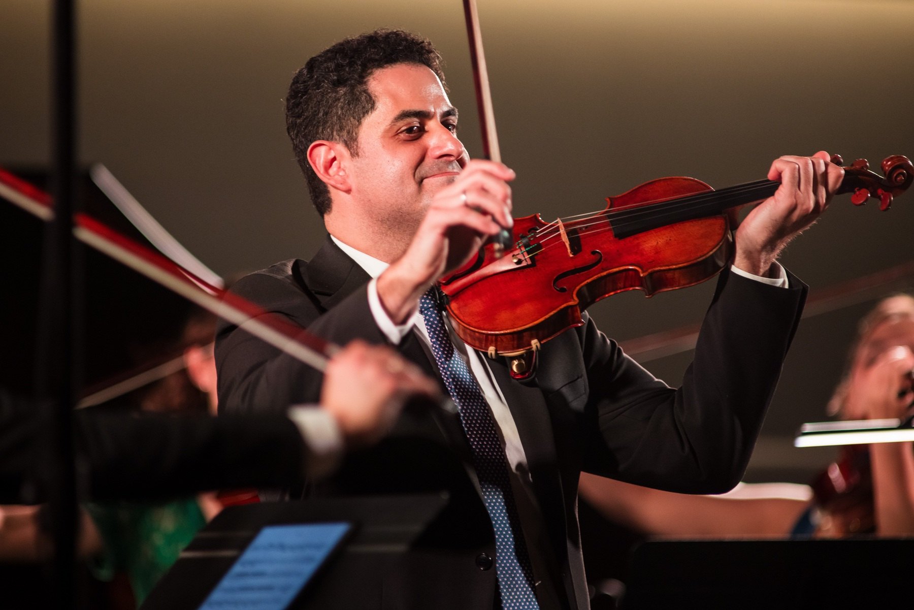 Arnaud Sussmann smiling as he plays the violin on stage with an ensemble, in warm light.