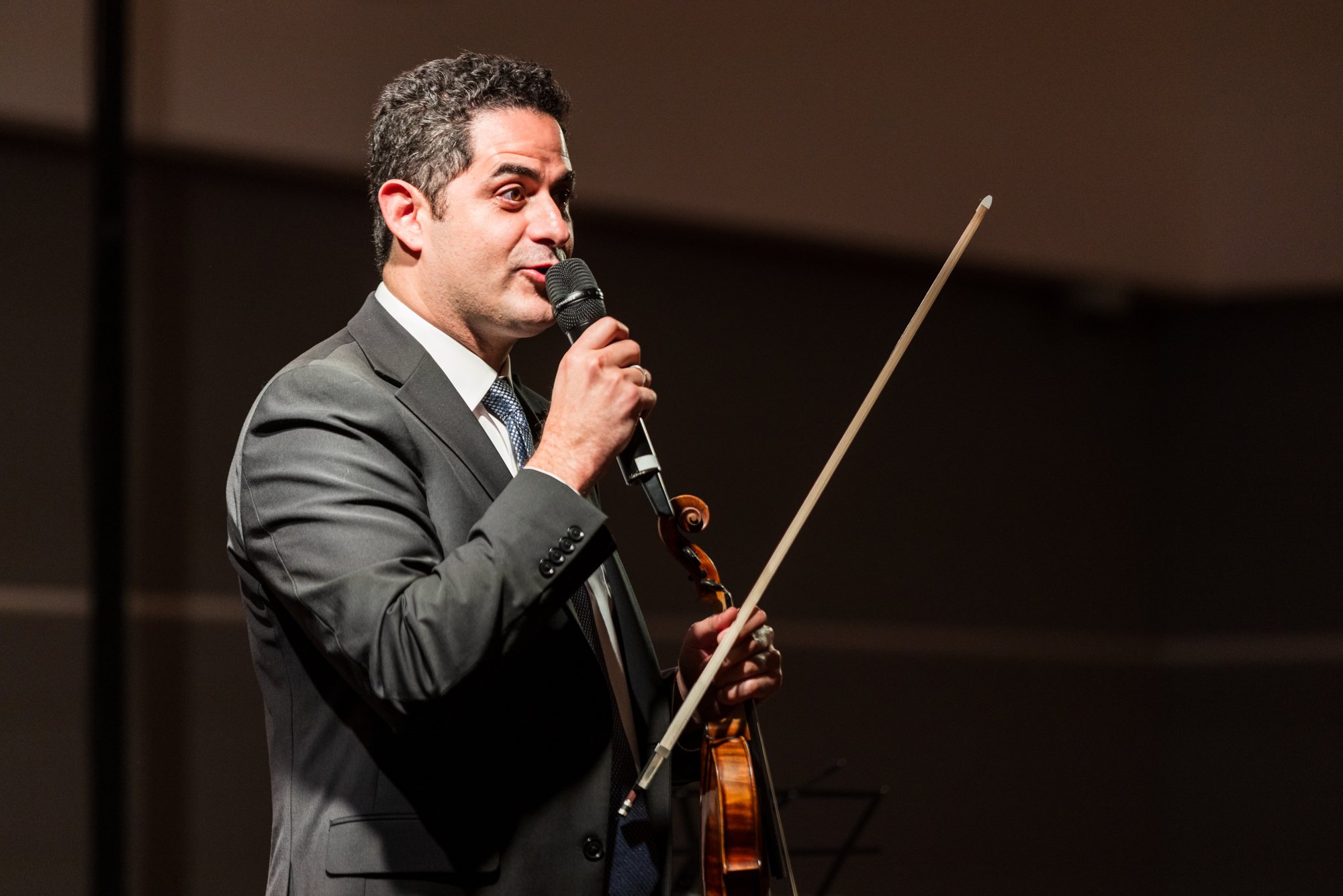 Arnaud Sussmann alone on stage, in a dark suit, holding a violin and bow and speaking into a microphone at The Lush Guitar, Chamber Music Society of Palm Beach.