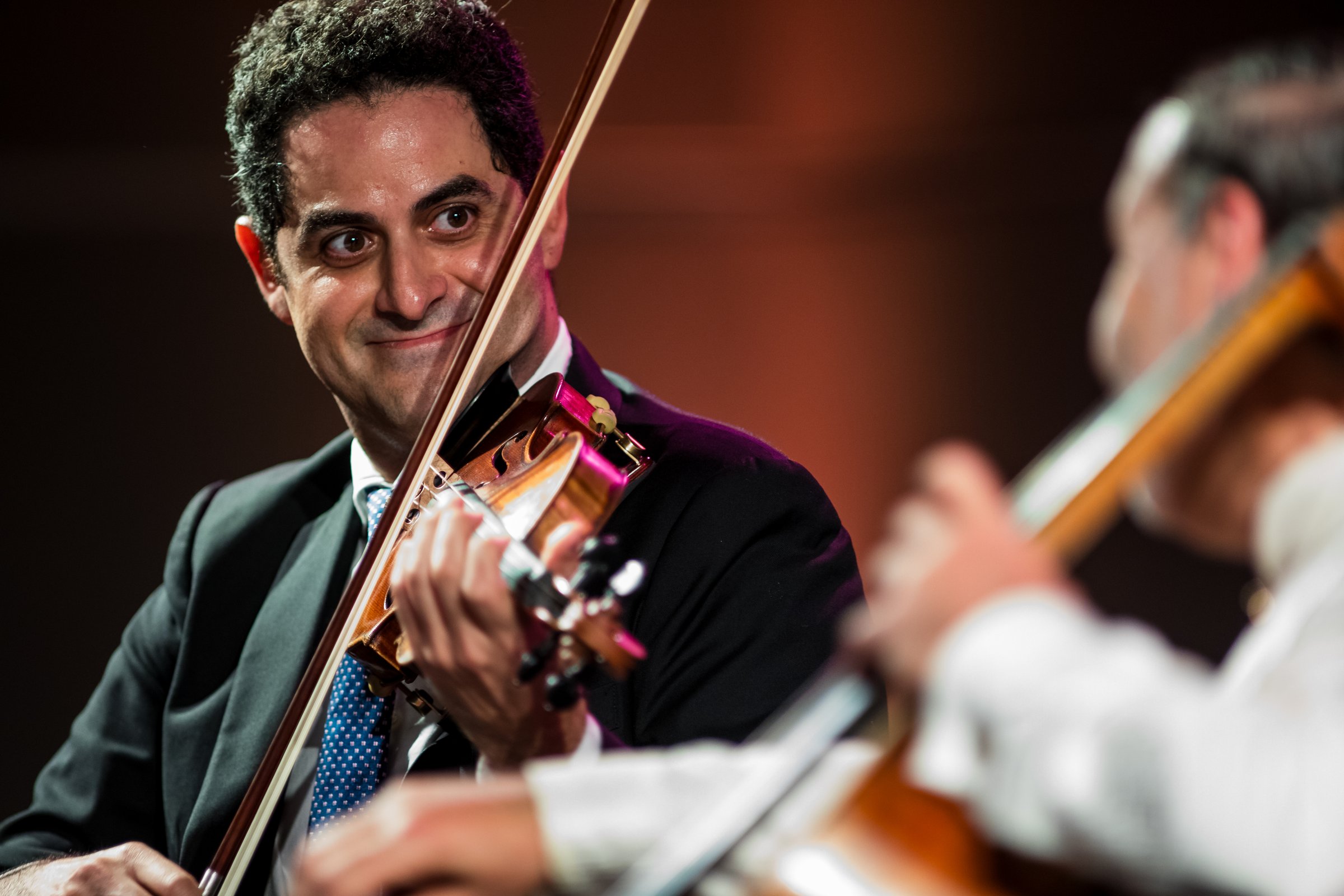 Arnaud Sussmann smiling on stage with his violin, looking sideways, with a blurred cellist in the right foreground.
