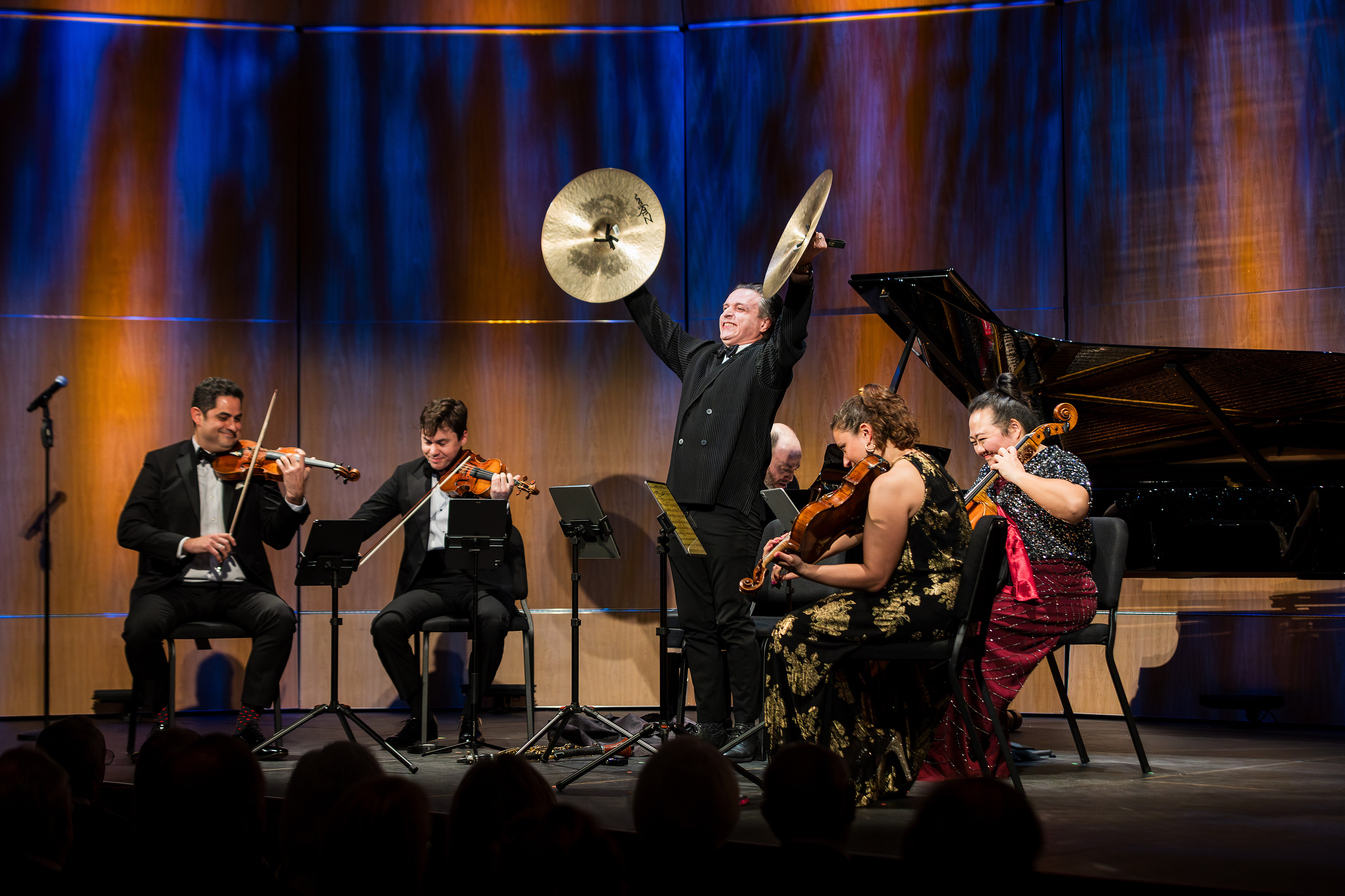 Chamber Music Society of Palm Beach gala: Arnaud Sussmann playing violin at left while the clarinetist raises crash cymbals overhead, with colleagues on a warmly lit stage.